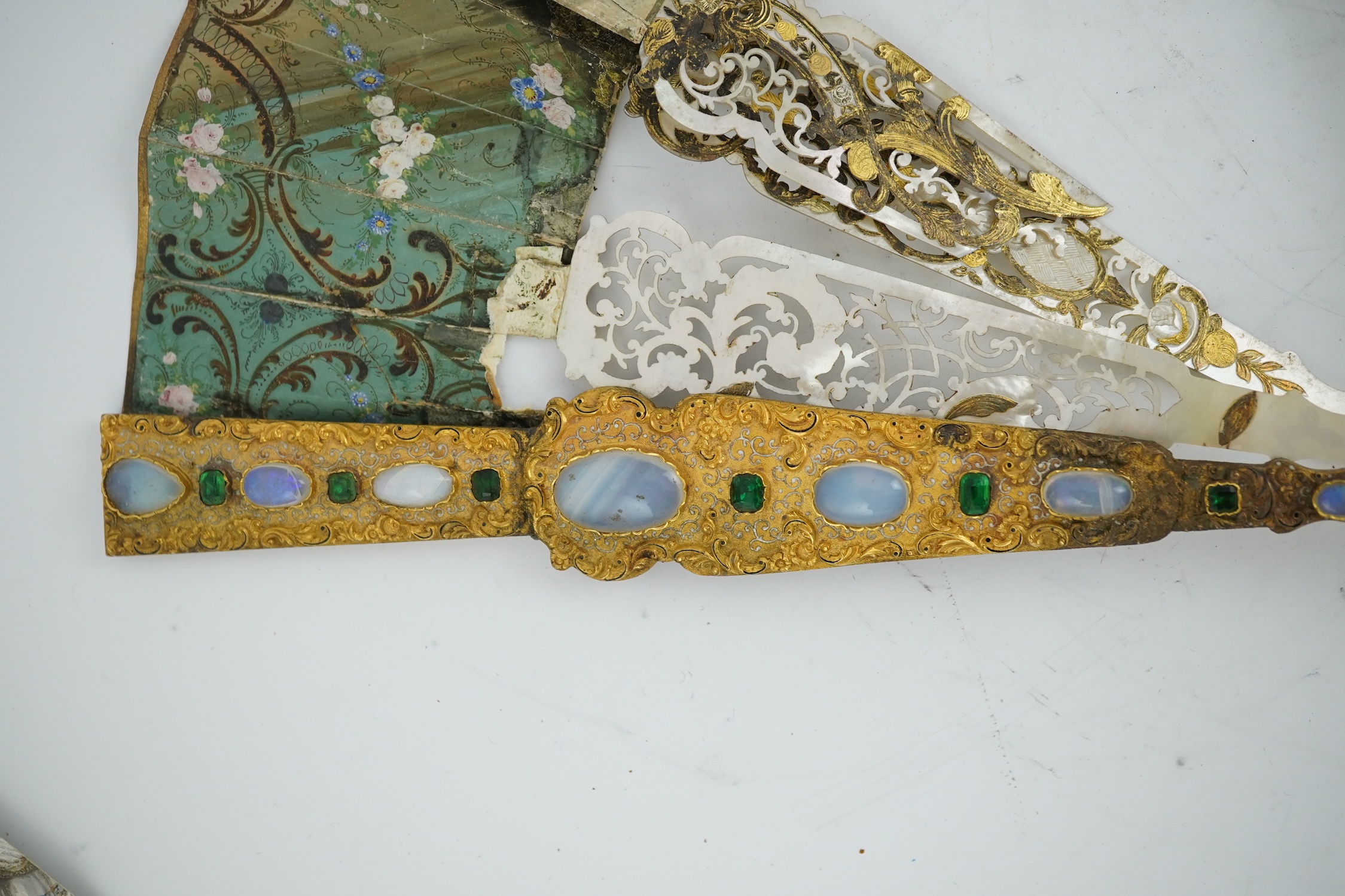 Two giltwood fans and display cases, one on a pedestal base housing an 18th century French figural hand painted fan with bone and gilt decorated guards and sticks, the other set with paste gilt metal guards on mother of